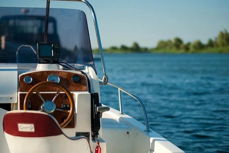 At the helm of a small boat on calm water, a tree line on the far shore