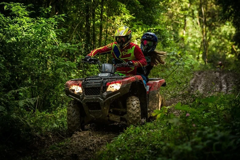 Two riders in helmets on an ATV on a muddy wooded trail